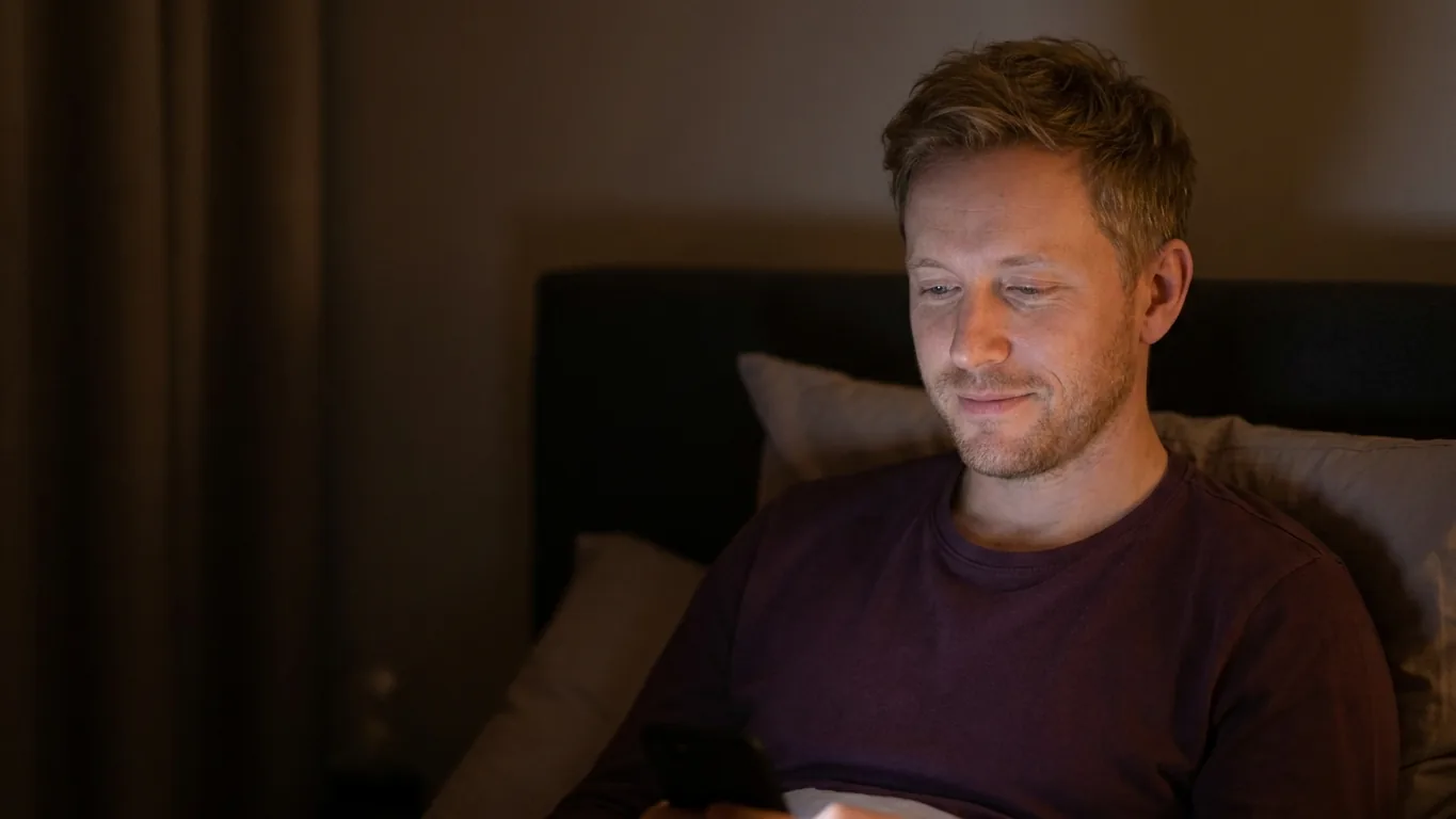 A man in bed at night, his face lit by the warm, low glow of his phone